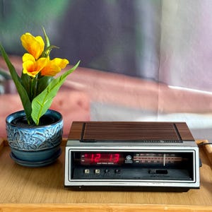 May include: A vintage General Electric electronic digital clock radio with a wood-grain top, a silver-toned front panel, and a red LED display showing 12:13. Beside it sits a blue ceramic planter with a textured pattern containing yellow flowers.