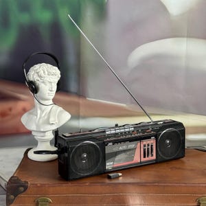 Könnte beinhalten: Ein schwarzer Vintage JVC RC-22-Boombox mit silberner Antenne, schwarzen Lautsprechern und einem rosa-schwarzen Bedienfeld. Links befindet sich eine weiße Büste einer klassischen Figur mit Kopfhörern. Die Boombox steht auf einem braunen Koffer.