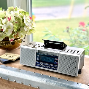 Könnte beinhalten: Ein silbernes und graues GE-Boombox mit offenem Kassettenrekorder. Die Digitalanzeige zeigt die Uhrzeit an. Das Radio verfügt über AM/FM-Einstellungen. Eine Kassette ist links sichtbar. Eine goldene Vase mit Blumen befindet sich im Hintergrund.