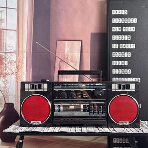 Puede incluir: Un radiocassette estéreo portátil Sanyo de color negro con dos prominentes rejillas de altavoz circulares rojas, una platina de casete central, un ecualizador gráfico de 3 bandas y una larga antena telescópica plateada.