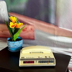 Puede incluir: Un radio reloj despertador vintage de color blanco hueso de General Electric con un auricular de teléfono encima. La pantalla digital muestra la hora 12:06 en rojo y el dial de radio muestra frecuencias AM y FM. Al lado hay una maceta azul con flores amarillas.
