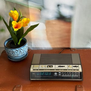 May include: A vintage General Electric digital clock radio with a dark grey plastic casing, a green LED display showing 12:03 PM, and a radio tuner dial. The device features various control buttons, sliders, and a speaker grille on the top surface.