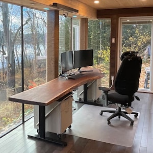 Puede incluir: Un gran escritorio de madera de borde vivo con un acabado de madera natural. El escritorio tiene un marco de metal negro y está sentado en un piso de madera oscura. Hay dos monitores de computadora en el escritorio, una silla de oficina negra y una ventana con vista a los árboles y un lago.