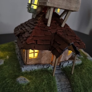 May include: A miniature model of a house with a brown shingled roof and a stone foundation. The house has a small porch and a window with a light inside. The house is sitting on a green grassy base with a stone path leading up to the porch.
