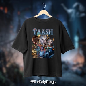 May include: Black t-shirt with the word "TAASH" in blue and white lettering. The shirt features a graphic of a person with silver hair and blue skin, surrounded by other characters. The Daily Things logo is at the bottom.