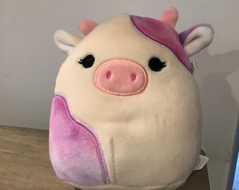 Peut inclure: Un jouet en peluche en forme de vache avec un design ombré blanc et violet. La vache a des yeux noirs, un nez rose et une bouche rose.