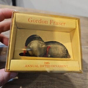 May include: A vintage 1985 Gordon Fraser annual dated ornament in a clear plastic box. The ornament is made of dark brown material and has a red string.