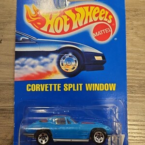 Op de afbeelding: Een blauwe Hot Wheels-speelgoedauto, een Corvette Split Window, in de originele verpakking. De auto staat op een blauwe achtergrond met het Hot Wheels-logo en de Mattel-branding.