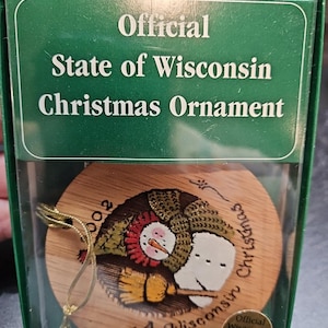 May include: A wooden Christmas ornament featuring a snowman holding a broom. The ornament is round and has a gold string attached. The text "A Wisconsin Christmas 2001" is written on the ornament. The ornament is in a green box with the text "Official State of Wisconsin Christmas Ornament".