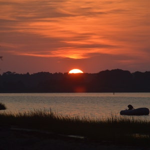 Op de afbeelding: Een silhouet van een kleine boot op een kalm wateroppervlak met een heldere oranje zonsondergang op de achtergrond.