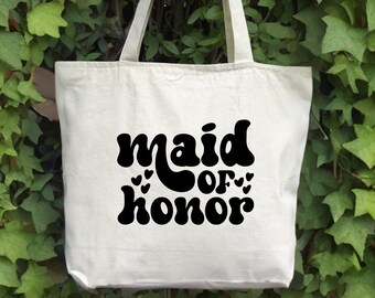 May include: A white canvas tote bag with black text that reads "maid of honor" in a retro font. The text is surrounded by small hearts.