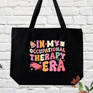 May include: Black canvas tote bag with the text "In My Occupational Therapy Era" in colorful retro font. The text is surrounded by small illustrations of occupational therapy tools.