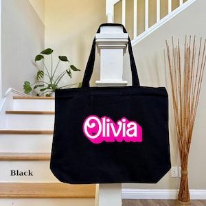 K&ouml;nnte beinhalten: Schwarze Stofftasche mit einem pinkfarbenen und wei&szlig;en "Olivia"-Design.