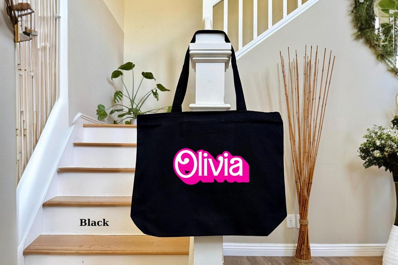 K&ouml;nnte beinhalten: Schwarze Stofftasche mit einem pinkfarbenen und wei&szlig;en "Olivia"-Design.