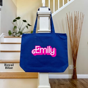 K&ouml;nnte beinhalten: Eine k&ouml;nigsblaue Tragetasche mit dem Namen "Emily" in pinken 3D-Buchstaben.