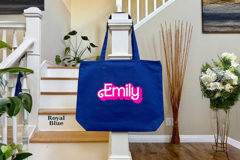 K&ouml;nnte beinhalten: Eine k&ouml;nigsblaue Tragetasche mit dem Namen "Emily" in pinken 3D-Buchstaben.