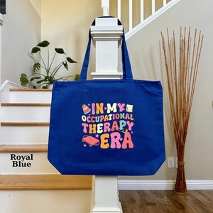 May include: A royal blue canvas tote bag with the text "In My Occupational Therapy Era" in colorful, retro-style lettering. The text is surrounded by stars and other small, colorful graphics.