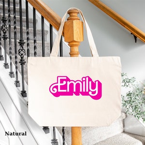 K&ouml;nnte beinhalten: Eine nat&uuml;rliche Canvas-Tragetasche mit dem Namen "Emily" in pinken Buchstaben.