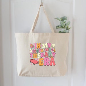 May include: A beige canvas tote bag with the text "In My Occupational Therapy Era" in colorful letters and illustrations.