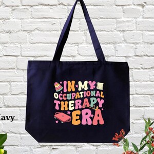 May include: A navy blue tote bag with the text "In My Occupational Therapy Era" in colorful, retro-style lettering. The text is surrounded by illustrations of occupational therapy tools, such as a stacking toy, a pencil, and a jump rope.