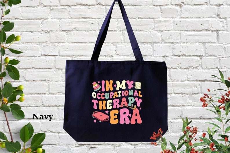 May include: A navy blue tote bag with the text "In My Occupational Therapy Era" in colorful, retro-style lettering. The text is surrounded by illustrations of occupational therapy tools, such as a stacking toy, a pencil, and a jump rope.