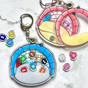 May include: Two clear acrylic shaker keychains shaped like igloos, one blue and one pink, containing small loose character charms. The blue igloo features a blue roof and interior scene with multiple colorful character inserts. The pink igloo has a pink roof and interior details. Several additional small character charms are scattered nearby.