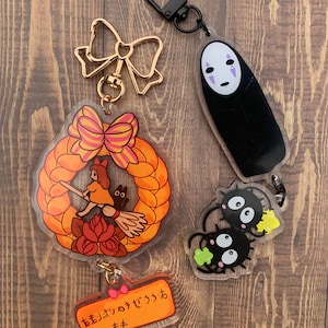 Pode incluir: Um conjunto de tr&ecirc;s chaveiros de acr&iacute;lico com personagens do filme de anima&ccedil;&atilde;o 'A Viagem de Chihiro'. O primeiro chaveiro apresenta uma coroa com uma menina em uma vassoura, o segundo chaveiro apresenta um personagem preto e branco com um rosto, e o terceiro chaveiro apresenta dois personagens preto e branco com olhos grandes. Os chaveiros s&atilde;o presos a an&eacute;is de metal dourado e preto.