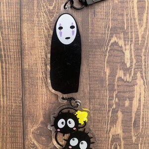 Pode incluir: Um chaveiro de acr&iacute;lico preto e branco com Sem Rosto do filme de anima&ccedil;&atilde;o A Viagem de Chihiro. O chaveiro tamb&eacute;m apresenta dois personagens pretos e brancos com detalhes amarelos e verdes.