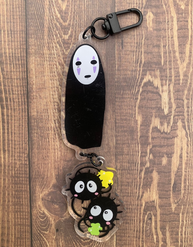 Pode incluir: Um chaveiro de acr&iacute;lico preto e branco com Sem Rosto do filme de anima&ccedil;&atilde;o A Viagem de Chihiro. O chaveiro tamb&eacute;m apresenta dois personagens pretos e brancos com detalhes amarelos e verdes.