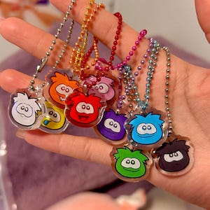 Puede incluir: Una colección de llaveros acrílicos coloridos con personajes de dibujos animados con diversas expresiones. Cada llavero tiene una cadena de bolas plateada. Los personajes son en tonos de blanco, naranja, amarillo, rojo, rosa, morado, azul, verde y negro.