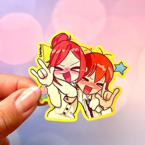 Puede incluir: Una pegatina amarilla con dos personajes de estilo anime con pelo rojo, sonriendo y haciendo gestos con las manos. La pegatina tiene un borde blanco y el texto "UwUmii". También es visible una estrella azul.
