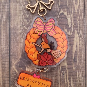 Pode incluir: Um chaveiro dourado com um amuleto transparente que apresenta uma ilustra&ccedil;&atilde;o de desenho animado de uma menina montando uma vassoura com um gato preto. O amuleto tem a forma de uma coroa com um la&ccedil;o rosa e amarelo. O chaveiro tamb&eacute;m inclui um amuleto transparente menor com o texto "おまいさんのデジっつおキキ" em laranja.