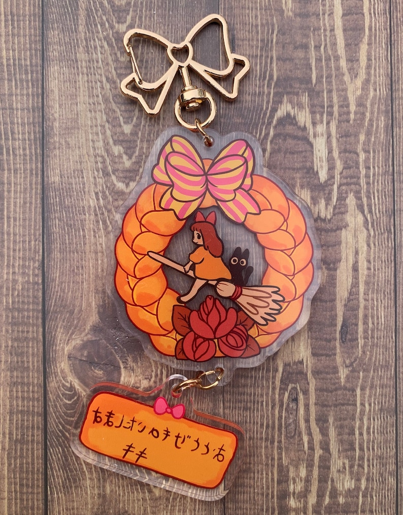 Pode incluir: Um chaveiro dourado com um amuleto transparente que apresenta uma ilustra&ccedil;&atilde;o de desenho animado de uma menina montando uma vassoura com um gato preto. O amuleto tem a forma de uma coroa com um la&ccedil;o rosa e amarelo. O chaveiro tamb&eacute;m inclui um amuleto transparente menor com o texto "おまいさんのデジっつおキキ" em laranja.