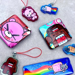 May include: A collection of glossy acrylic charms and pins featuring cartoon characters including a pink bear, a brown square creature, and a gray cat with a rainbow trail. The items include various shapes like squares, rectangles, and custom die-cut outlines.