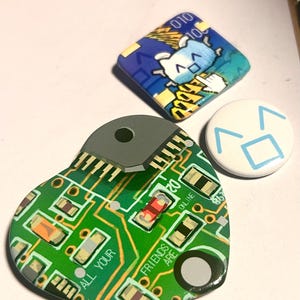 May include: Three pin-back buttons: a heart-shaped button with a green circuit board pattern and the text ALL YOUR FRIENDS ARE ONLINE, a square button with a blue pixelated character, and a small round white button with a blue geometric face.