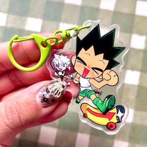 May include: A clear acrylic keychain with a cartoon character on a skateboard, and a green clip. The character has black spiky hair, a white shirt, and green shorts and socks. Another character is also visible.