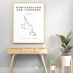May include: A black and white line art map of Newfoundland and Labrador, Canada. The map is framed in a light wood frame and is hanging on a light gray wall. The coordinates 53.1355° N, 57.6604° W are printed below the map.