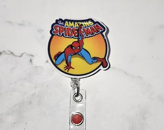 May include: A red and blue cartoon image of Spiderman with the text "the AMAZING SPIDER-MAN" on a yellow background. The image is attached to a clear plastic retractable badge holder with a red button.