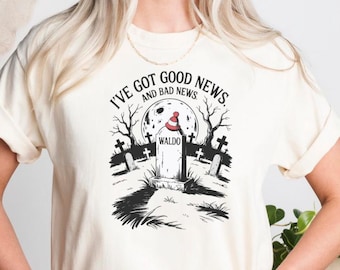 Może przedstawiać: Kremowa koszulka z napisem "I've got good news and bad news". Grafika przedstawia nagrobek z napisem "Waldo" i upiorną scenę. Modelka ma na sobie niebieskie dżinsy.