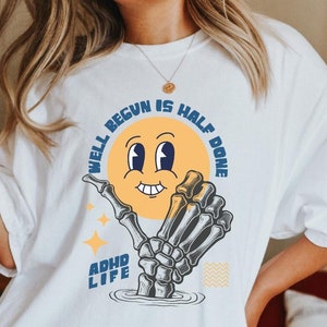 Könnte beinhalten: Weißes T-Shirt mit einem Grafikdruck eines lächelnden Gesichts, das von einer Skeletthand gehalten wird. Der Text auf dem Shirt lautet "Well begun is half done" und "ADHD Life".