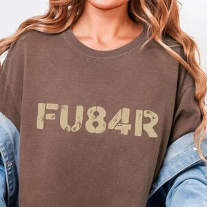 Puede incluir: Camiseta color espresso con el texto "FU84R" en una fuente desgastada de color beige. La camiseta forma parte de la colección Xanadu Hills. La modelo lleva una chaqueta vaquera.