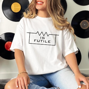 May include: White t-shirt with the text "IS FUTILE" in a black box with a circuit diagram above it. The shirt is worn by a person. The background includes black vinyl records.