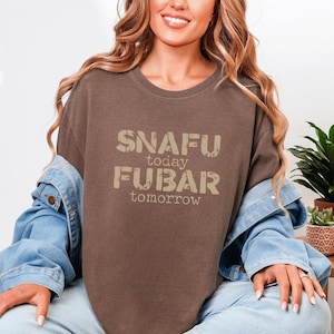Puede incluir: Camiseta color espresso con el texto "SNAFU today FUBAR tomorrow" en una fuente desgastada. La camiseta es parte de la colección Xanadu Hills. La modelo lleva una chaqueta vaquera.