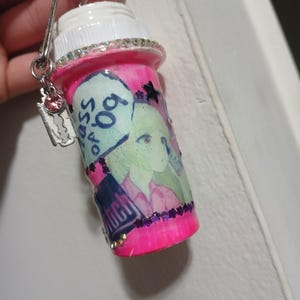 May include: A bright pink cylindrical container with a white screw-top lid, decorated with a cartoon character illustration, a speech bubble reading "class of 09", and a "bitch" label. It features a silver keychain with a black butterfly and razor charm.