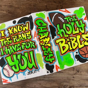 May include: Three canvases with colorful graffiti-style lettering. The first canvas says "I know the plans I have for you" with a baseball and the scripture "JER 29:11". The second canvas says "God is my everything" with a basketball. The third canvas says "The Holy Bible" with a football and a basketball.