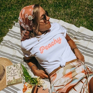 May include: A woman wearing a white t-shirt with the word "Peachy" in pink lettering, a brown and white bandana, and a patterned skirt. She is lying on a striped blanket with a book and a hat beside her.