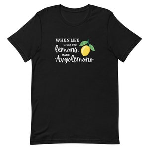Puede incluir: Una camiseta negra de manga corta con un estampado gr&aacute;fico que incluye el texto "WHEN LIFE GIVES YOU lemons, MAKE Avgolemono" en letras blancas y cursivas, acompa&ntilde;ado de una ilustraci&oacute;n de un lim&oacute;n amarillo con dos hojas verdes.