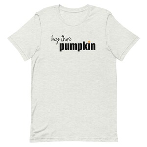 Pode incluir: Camiseta cinza claro com a frase "hey there pumpkin" em letras pretas e em bloco. Um pequeno gr&aacute;fico de ab&oacute;bora laranja est&aacute; acima da palavra "pumpkin". A camiseta &eacute; de gola redonda e manga curta.