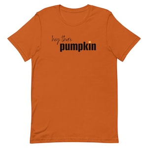Pode incluir: Uma camiseta laranja com a frase "hey there pumpkin" impressa em preto e letras de bloco. Um pequeno ponto laranja est&aacute; acima da palavra "pumpkin". A camisa &eacute; feita de um material macio.