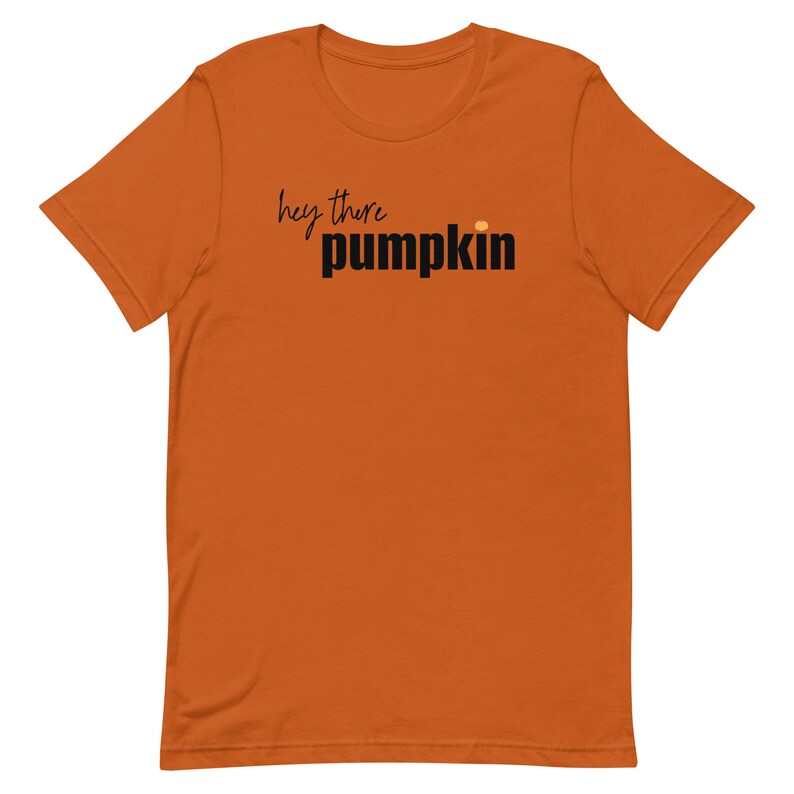 Pode incluir: Uma camiseta laranja com a frase "hey there pumpkin" impressa em preto e letras de bloco. Um pequeno ponto laranja est&aacute; acima da palavra "pumpkin". A camisa &eacute; feita de um material macio.